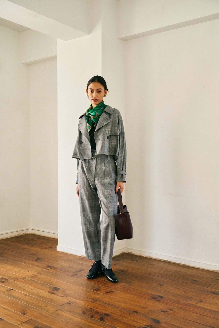 A person with short blond hair wearing oversized black sunglasses, a Marie Cropped Glen Plaid Jacket by Maison Lilli in a soft wool blend with an adjustable back waist, a beige shirt, and a gold chain necklace stands against a neutral gray background. The individual exudes a confident and stylish demeanor.