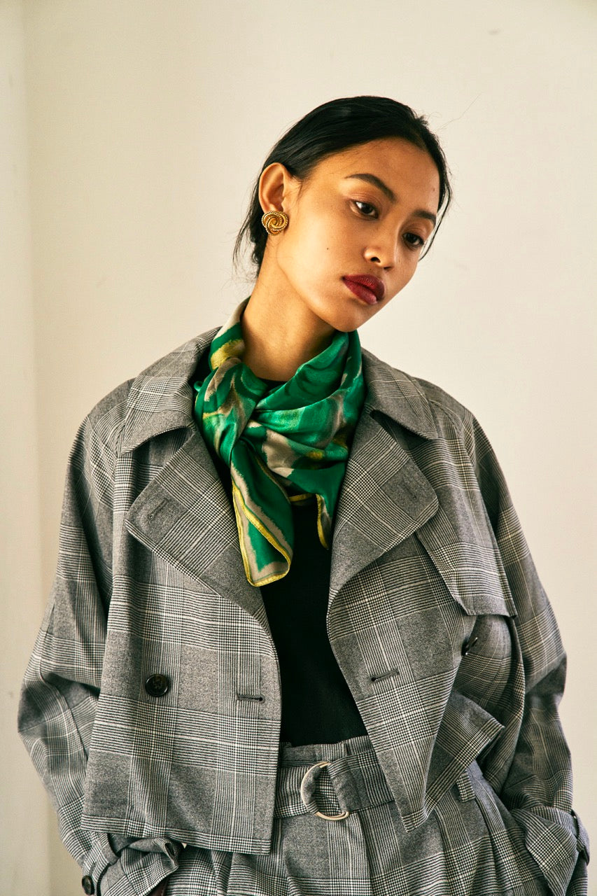 A person with short blond hair wearing oversized black sunglasses, a Marie Cropped Glen Plaid Jacket by Maison Lilli in a soft wool blend with an adjustable back waist, a beige shirt, and a gold chain necklace stands against a neutral gray background. The individual exudes a confident and stylish demeanor.