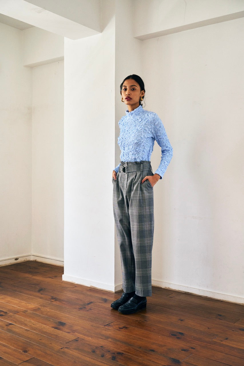 A person with short blond hair wearing oversized black sunglasses, a Marie Cropped Glen Plaid Jacket by Maison Lilli in a soft wool blend with an adjustable back waist, a beige shirt, and a gold chain necklace stands against a neutral gray background. The individual exudes a confident and stylish demeanor.