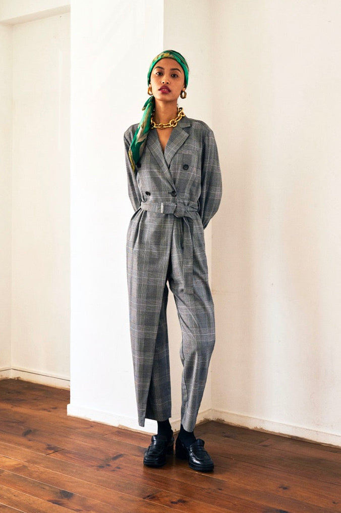A woman stands against a plain background wearing a Chloe Glen Plaid Tailored jumpsuit by Maison Lilli. She accessorizes with large black sunglasses, gold hoop earrings, and a gold necklace. Her hair is styled in an updo.