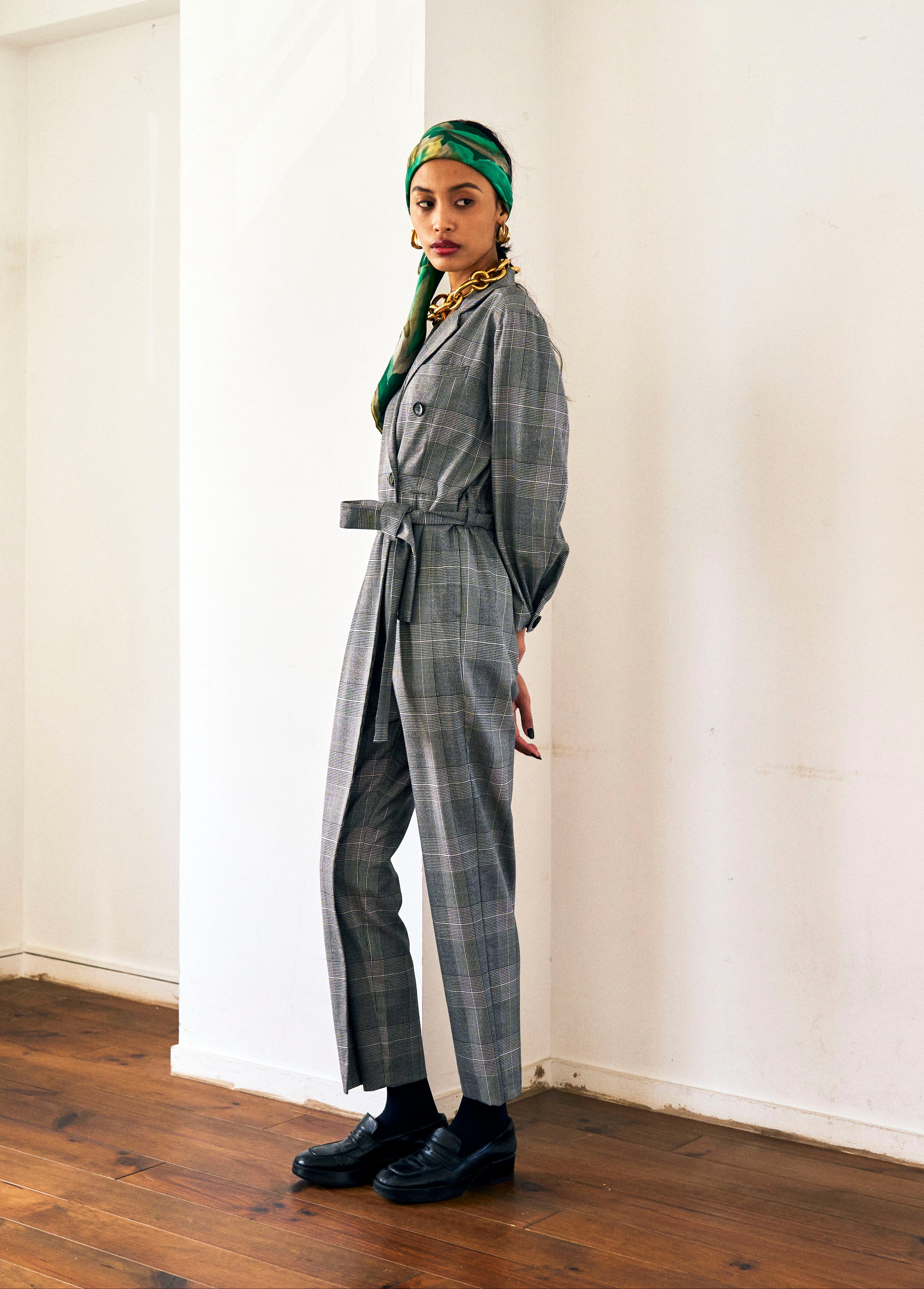 A woman stands against a plain background wearing a Chloe Glen Plaid Tailored jumpsuit by Maison Lilli. She accessorizes with large black sunglasses, gold hoop earrings, and a gold necklace. Her hair is styled in an updo.