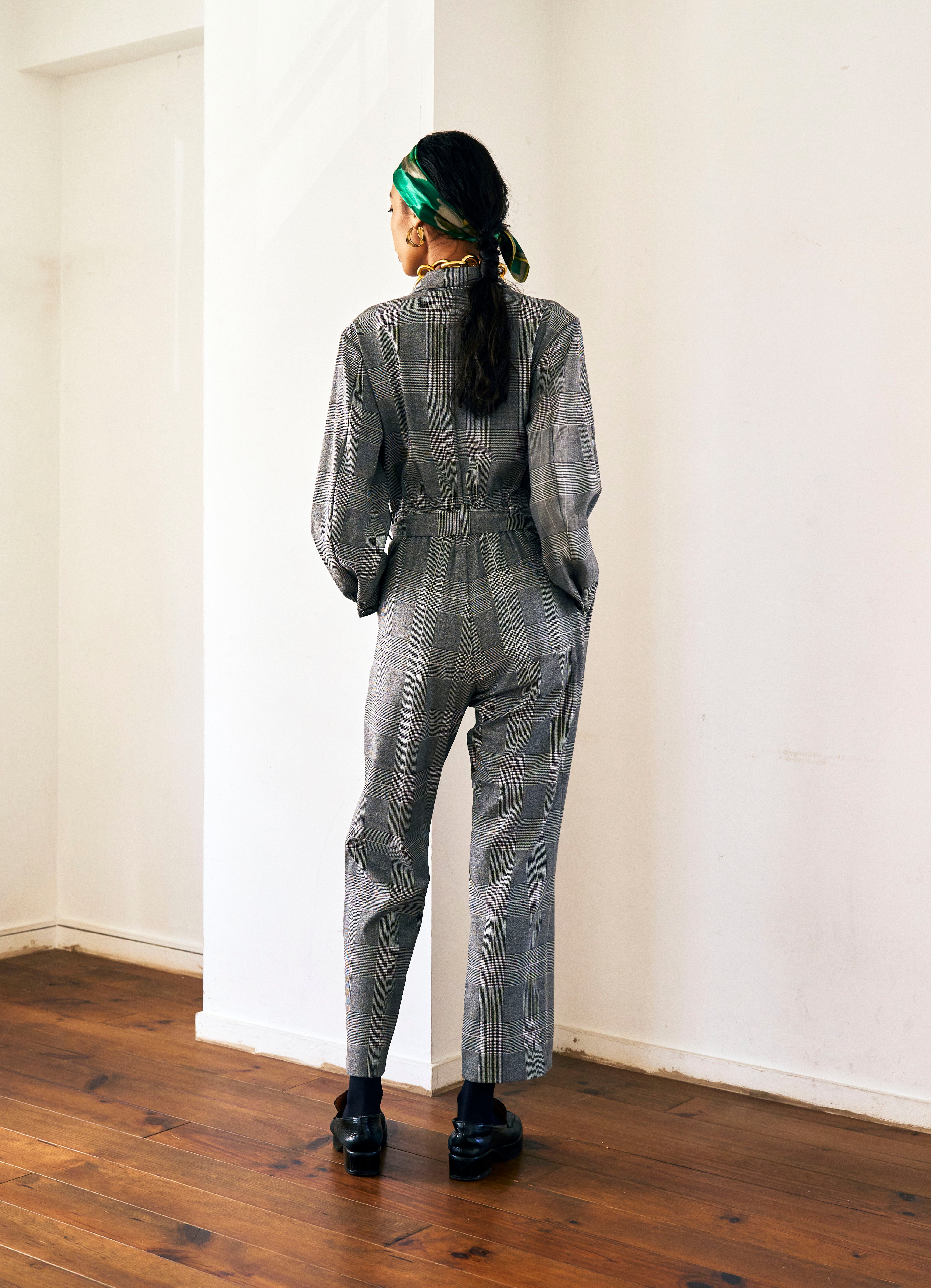 A woman stands against a plain background wearing a Chloe Glen Plaid Tailored jumpsuit by Maison Lilli. She accessorizes with large black sunglasses, gold hoop earrings, and a gold necklace. Her hair is styled in an updo.
