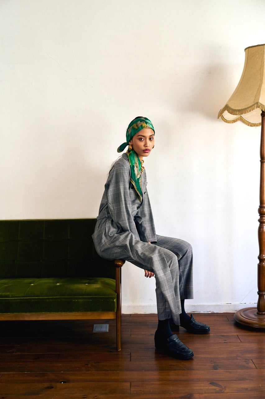 A woman stands against a plain background wearing a Chloe Glen Plaid Tailored jumpsuit by Maison Lilli. She accessorizes with large black sunglasses, gold hoop earrings, and a gold necklace. Her hair is styled in an updo.