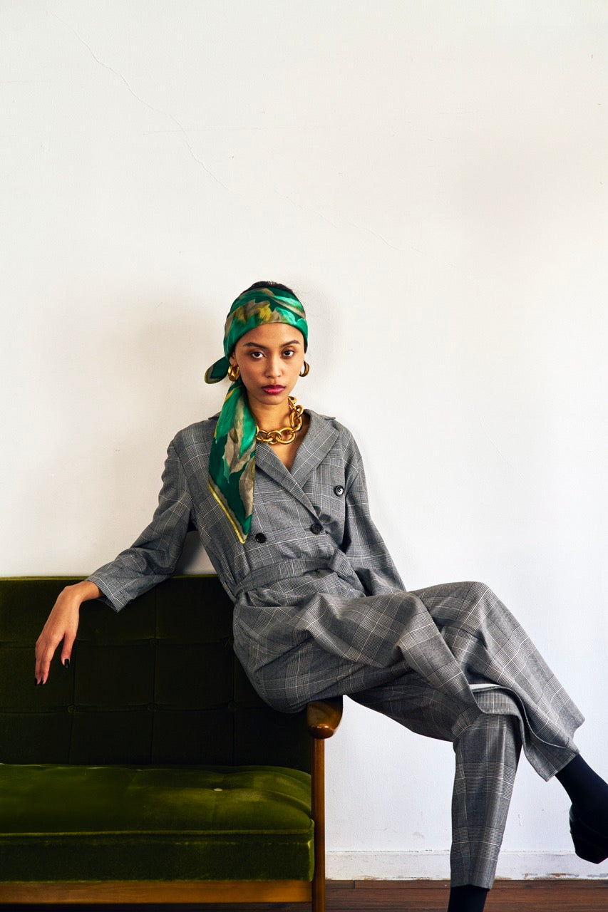 A woman stands against a plain background wearing a Chloe Glen Plaid Tailored jumpsuit by Maison Lilli. She accessorizes with large black sunglasses, gold hoop earrings, and a gold necklace. Her hair is styled in an updo.