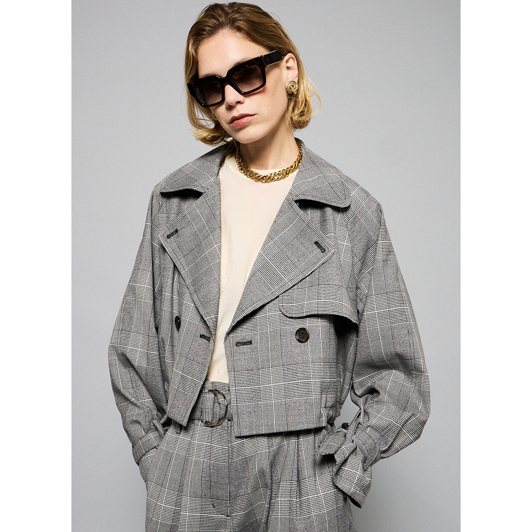 A person with short, blonde hair wearing large, dark sunglasses, a light chain necklace, and a chic Marie Cropped Glen Plaid Jacket by Maison Lilli over a light-colored shirt. The flattering silhouette exudes a stylish and modern vibe against a plain gray background.