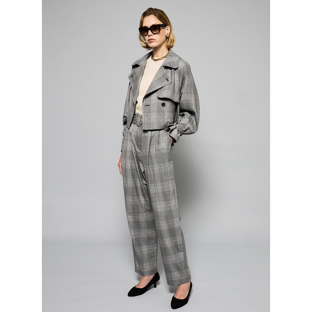 A person is wearing high-waisted, gray wool-blend Paperbag Pleated Pants - Glen Plaid by Maison Lilli with a large belt buckle. They have one hand in their pocket and are also wearing a loose-fitting short-sleeved off-white top and black heels. The background is plain light gray.