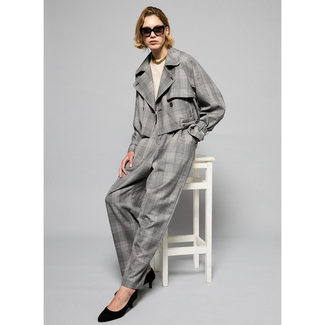 A person with short, blonde hair wearing large, dark sunglasses, a light chain necklace, and a chic Marie Cropped Glen Plaid Jacket by Maison Lilli over a light-colored shirt. The flattering silhouette exudes a stylish and modern vibe against a plain gray background.