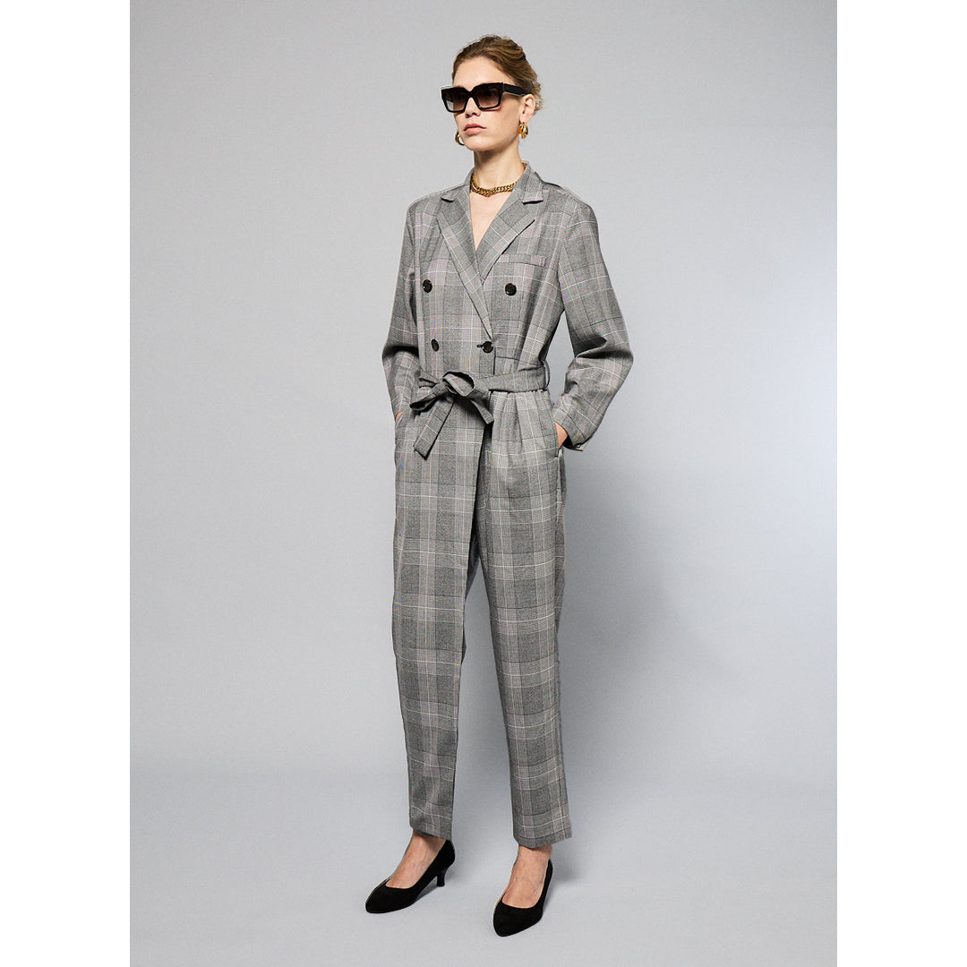 A woman stands against a plain background wearing a Chloe Glen Plaid Tailored jumpsuit by Maison Lilli. She accessorizes with large black sunglasses, gold hoop earrings, and a gold necklace. Her hair is styled in an updo.