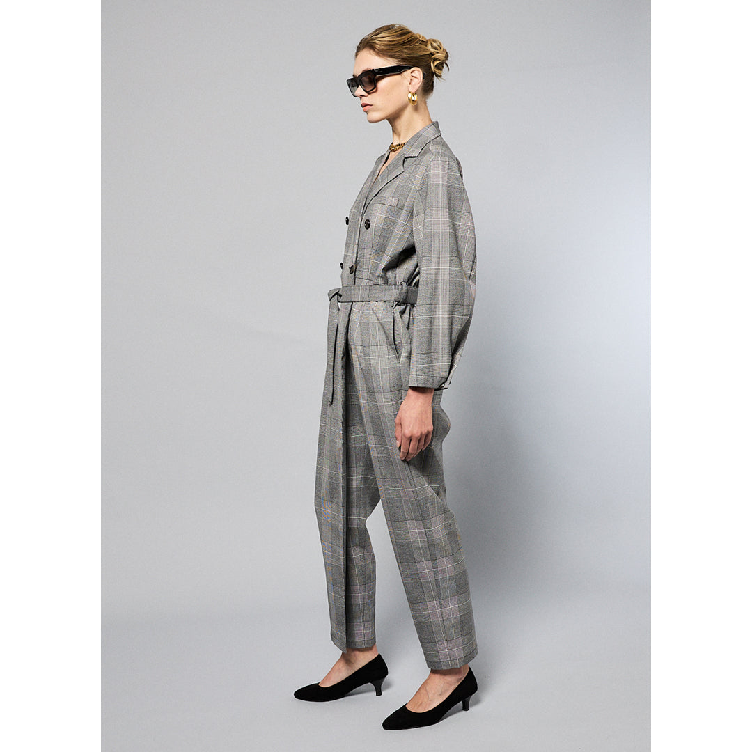 A woman stands against a plain background wearing a Chloe Glen Plaid Tailored jumpsuit by Maison Lilli. She accessorizes with large black sunglasses, gold hoop earrings, and a gold necklace. Her hair is styled in an updo.