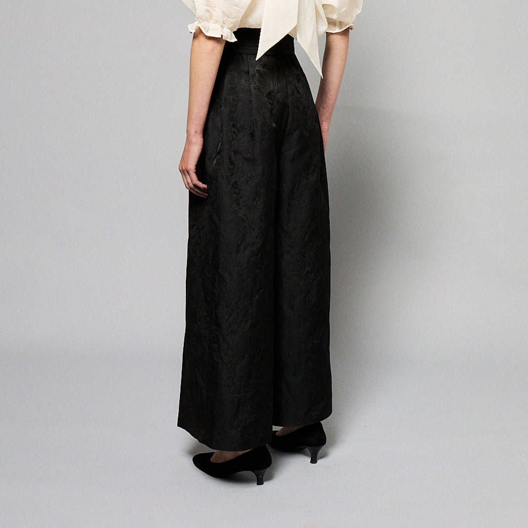 woman stands against a plain gray background, wearing a black, textured two-piece outfit with wide-leg, high-waisted Katia Pleated Hakama Pants by Maison Lilli and a short-sleeve, cropped blazer. She has her hair up and is wearing black high heels and drop earrings. A detachable belt cinches her waist.