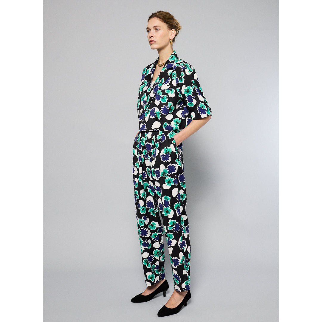 A person is wearing Maison Lilli Paperbag Pleated Pants - Floral Print with large blue, green, and white flowers. The pants have pockets and a removable belt. The person also sports a matching top and black high-heeled shoes against a plain light gray background.