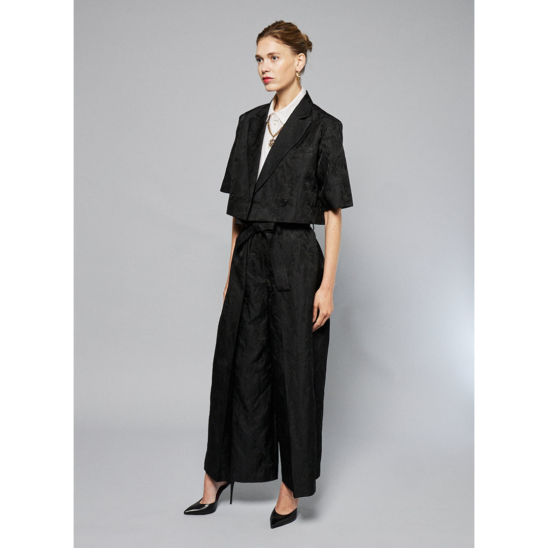 A person stands against a plain background, wearing a black Jacquard Short Sleeve Cropped Jacket by Maison Lilli. She also wear a white top underneath, gold hoop earrings, and a thick gold chain necklace.