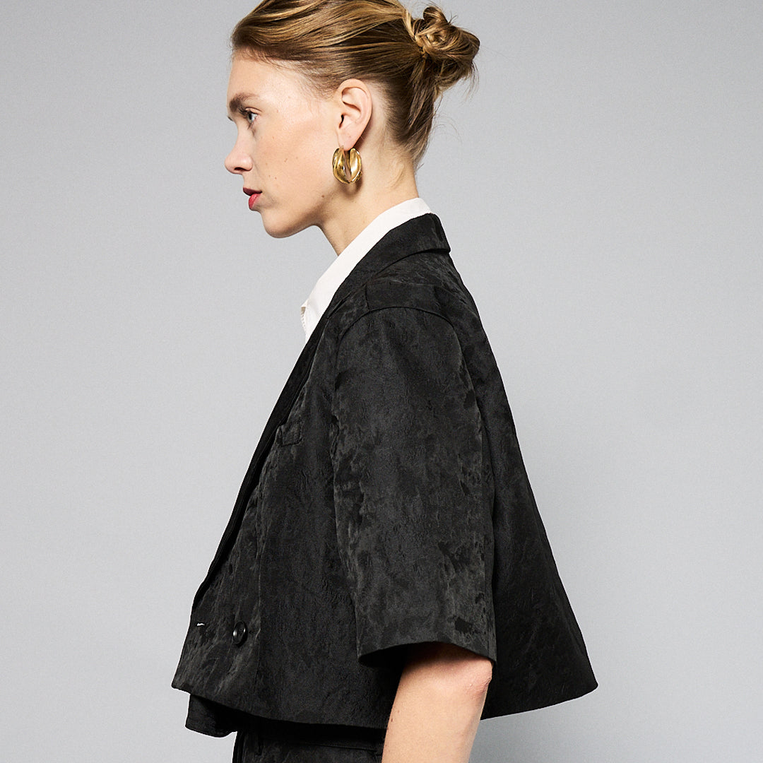 A person stands against a plain background, wearing a black Jacquard Short Sleeve Cropped Jacket by Maison Lilli. She also wear a white top underneath, gold hoop earrings, and a thick gold chain necklace.