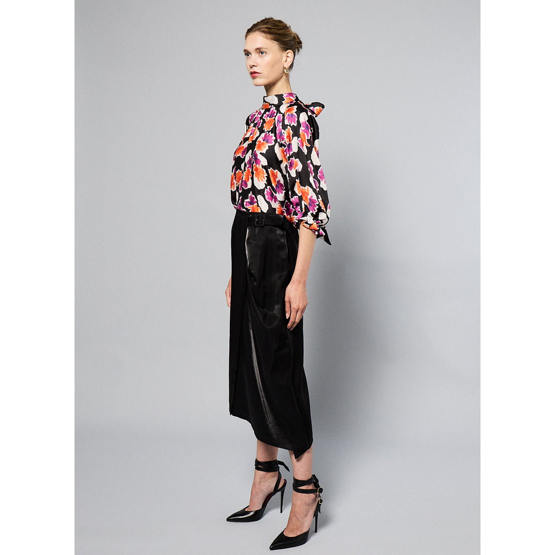 A person standing in front of a plain gray background, wearing a colorful floral blouse, a Maison Lilli Jodie Sparkling Satin Asymmetrical Draped Skirt, and black strappy high heels.