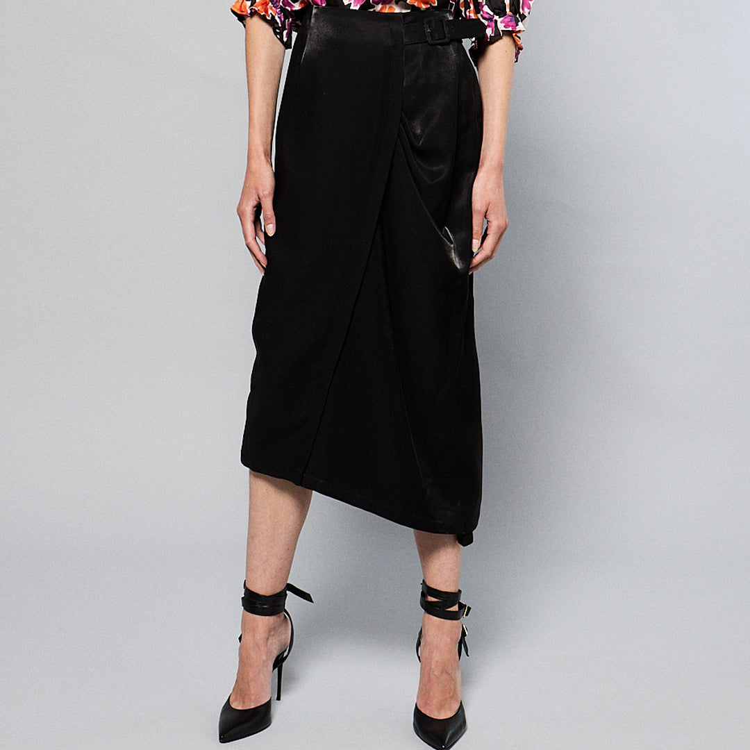 A person standing in front of a plain gray background, wearing a colorful floral blouse, a Maison Lilli Jodie Sparkling Satin Asymmetrical Draped Skirt, and black strappy high heels. The shot captures their lower torso and legs.