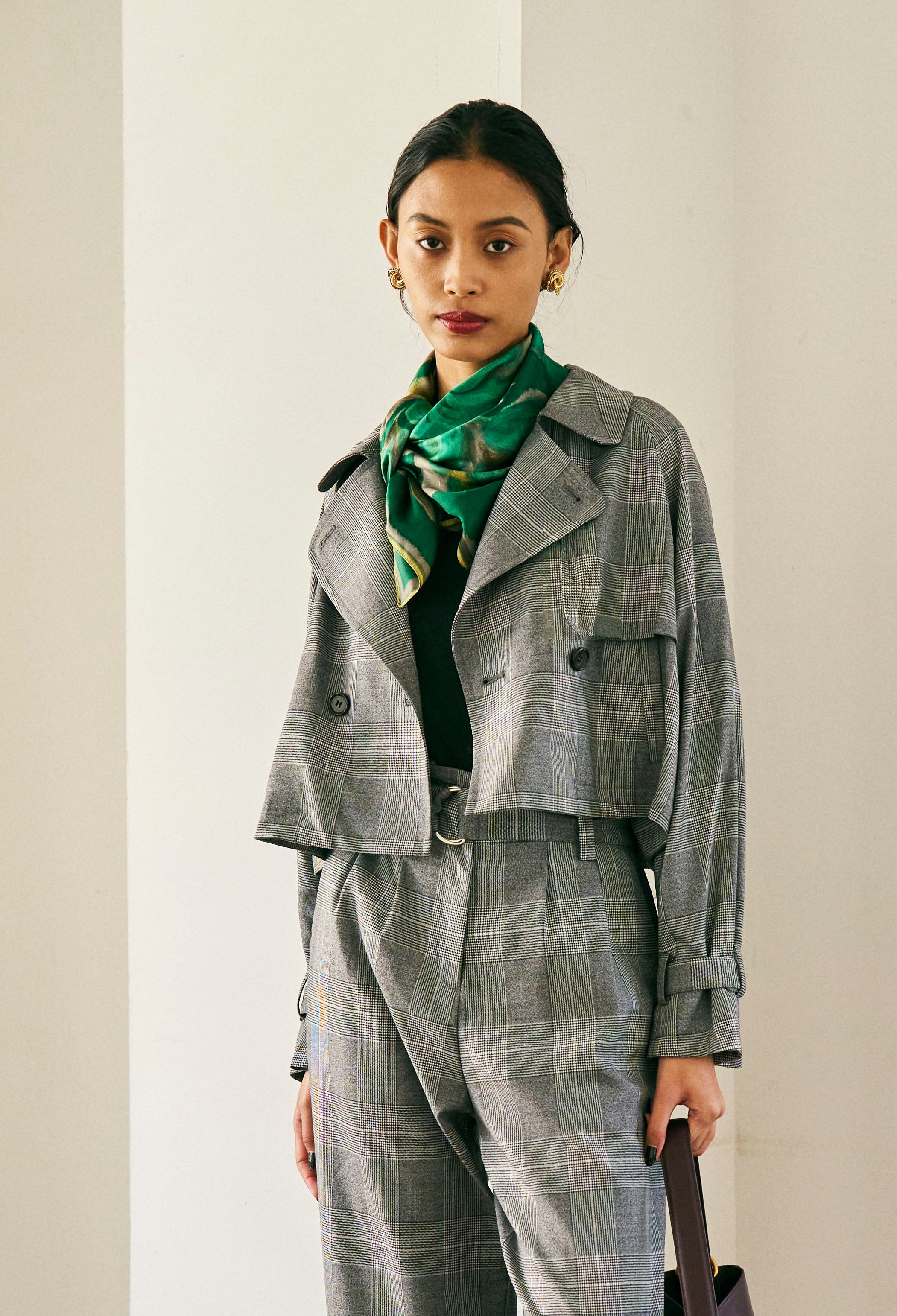 A person with short blond hair wearing oversized black sunglasses, a Marie Cropped Glen Plaid Jacket by Maison Lilli in a soft wool blend with an adjustable back waist, a beige shirt, and a gold chain necklace stands against a neutral gray background. The individual exudes a confident and stylish demeanor.