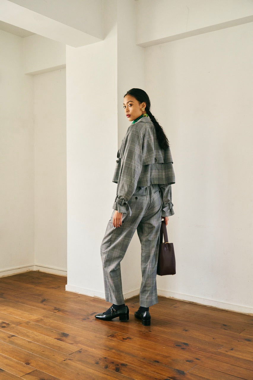 A person with short blond hair wearing oversized black sunglasses, a Marie Cropped Glen Plaid Jacket by Maison Lilli in a soft wool blend with an adjustable back waist, a beige shirt, and a gold chain necklace stands against a neutral gray background. The individual exudes a confident and stylish demeanor.