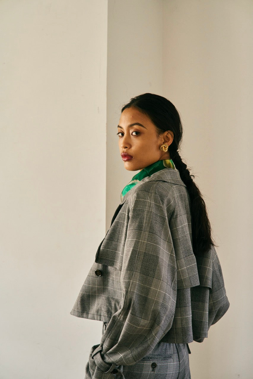 A person with short blond hair wearing oversized black sunglasses, a Marie Cropped Glen Plaid Jacket by Maison Lilli in a soft wool blend with an adjustable back waist, a beige shirt, and a gold chain necklace stands against a neutral gray background. The individual exudes a confident and stylish demeanor.