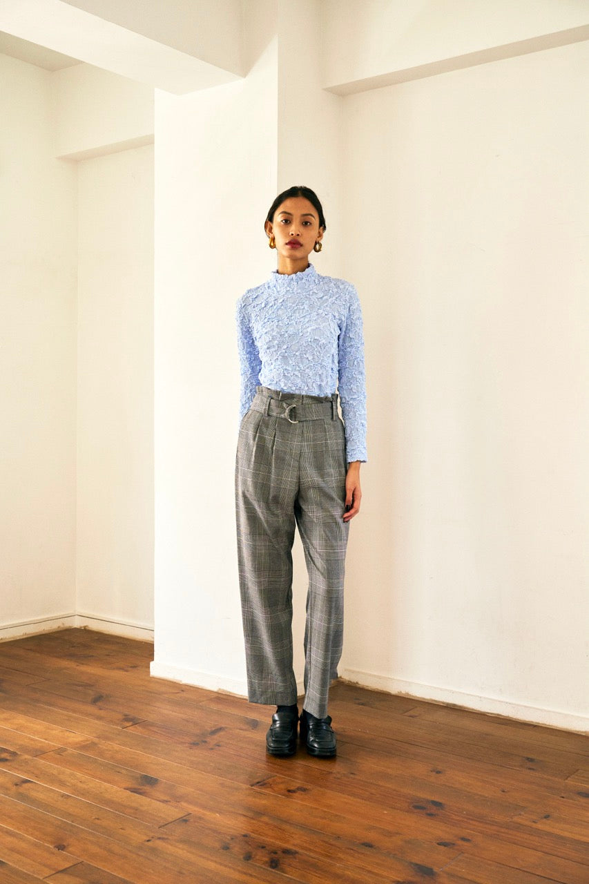 A person with short blond hair wearing oversized black sunglasses, a Marie Cropped Glen Plaid Jacket by Maison Lilli in a soft wool blend with an adjustable back waist, a beige shirt, and a gold chain necklace stands against a neutral gray background. The individual exudes a confident and stylish demeanor.