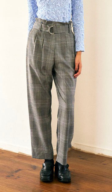 A person with short blond hair wearing oversized black sunglasses, a Marie Cropped Glen Plaid Jacket by Maison Lilli in a soft wool blend with an adjustable back waist, a beige shirt, and a gold chain necklace stands against a neutral gray background. The individual exudes a confident and stylish demeanor.