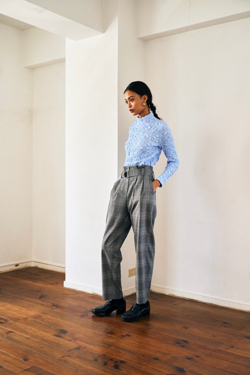 A person with short blond hair wearing oversized black sunglasses, a Marie Cropped Glen Plaid Jacket by Maison Lilli in a soft wool blend with an adjustable back waist, a beige shirt, and a gold chain necklace stands against a neutral gray background. The individual exudes a confident and stylish demeanor.
