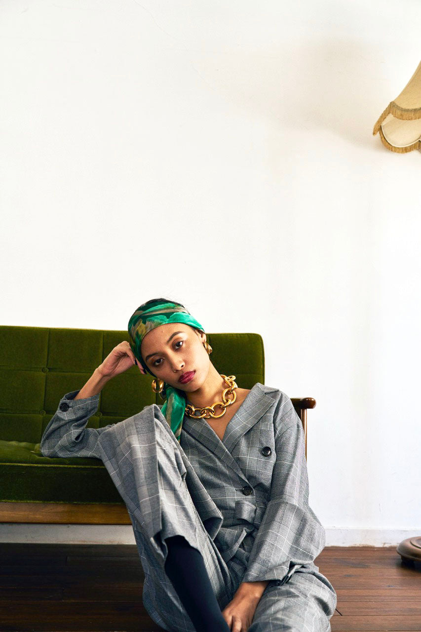 A woman stands against a plain background wearing a Chloe Glen Plaid Tailored jumpsuit by Maison Lilli. She accessorizes with large black sunglasses, gold hoop earrings, and a gold necklace. Her hair is styled in an updo.