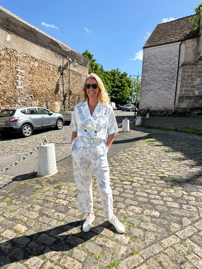 A person stands against a plain background, wearing a Marble Print Short Sleeve Cropped Jacket by Maison Lilli. She also wear a gold hoop earrings, and a thick gold chain necklace.