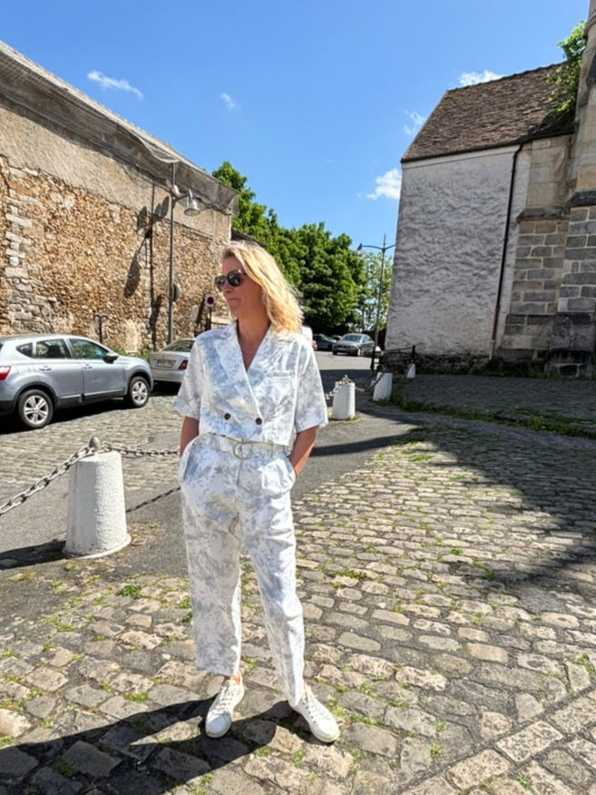 A person stands against a plain background, wearing a Marble Print Short Sleeve Cropped Jacket by Maison Lilli. She also wear a gold hoop earrings, and a thick gold chain necklace.