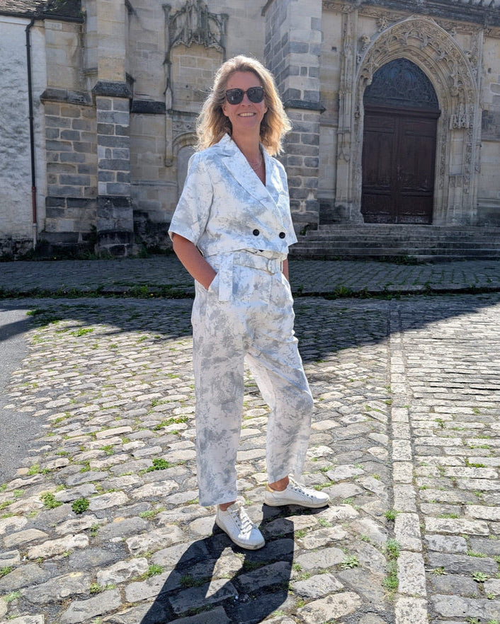 A person stands against a plain background, wearing a Marble Print Short Sleeve Cropped Jacket by Maison Lilli. She also wear a gold hoop earrings, and a thick gold chain necklace.