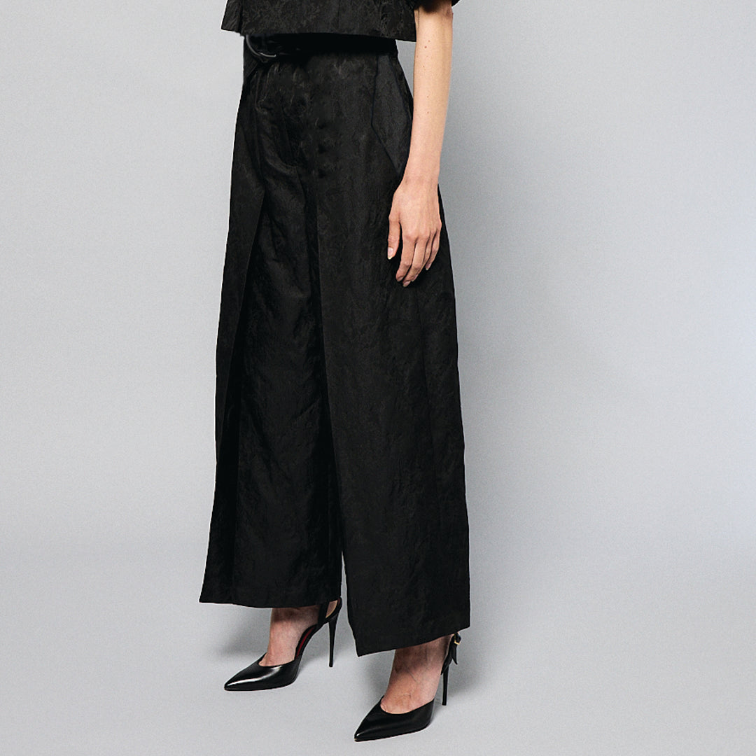  woman stands against a plain gray background, wearing a black, textured two-piece outfit with wide-leg, high-waisted Katia Pleated Hakama Pants by Maison Lilli and a short-sleeve, cropped blazer. She has her hair up and is wearing black high heels and drop earrings. A detachable belt cinches her waist.