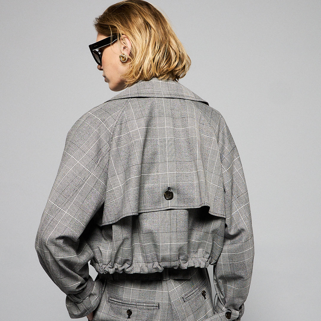 A person with short, blonde hair wearing large, dark sunglasses, a light chain necklace, and a chic Marie Cropped Glen Plaid Jacket by Maison Lilli over a light-colored shirt. The flattering silhouette exudes a stylish and modern vibe against a plain gray background.