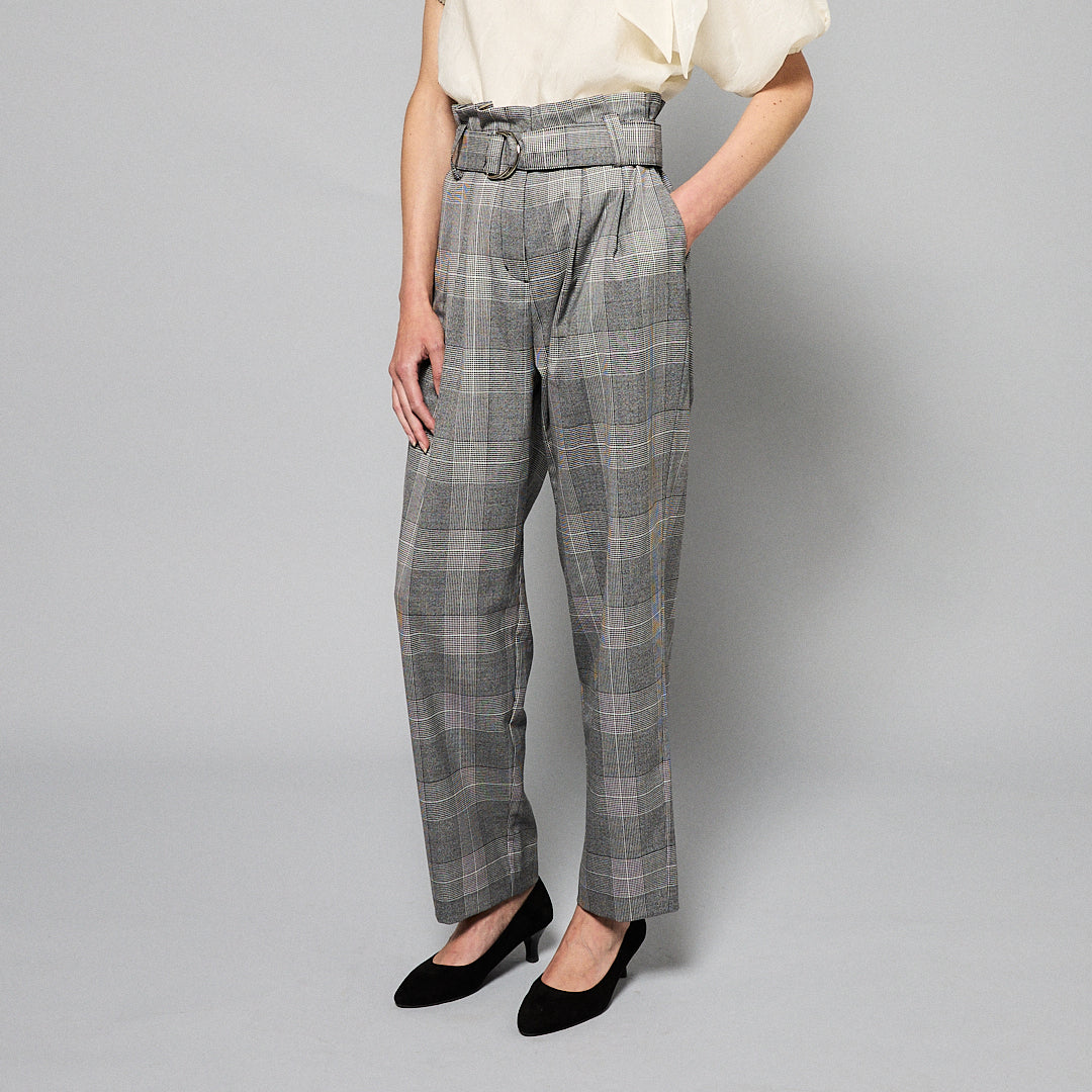 A person is wearing high-waisted, gray wool-blend Paperbag Pleated Pants - Glen Plaid by Maison Lilli with a large belt buckle. They have one hand in their pocket and are also wearing a loose-fitting short-sleeved off-white top and black heels. The background is plain light gray.