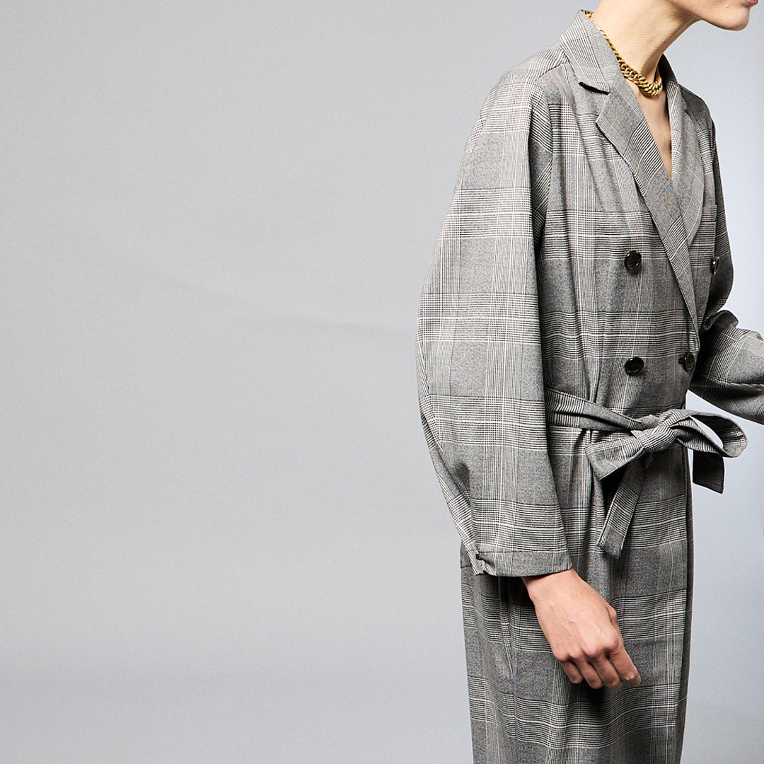 A woman stands against a plain background wearing a Chloe Glen Plaid Tailored jumpsuit by Maison Lilli. She accessorizes with large black sunglasses, gold hoop earrings, and a gold necklace. Her hair is styled in an updo.
