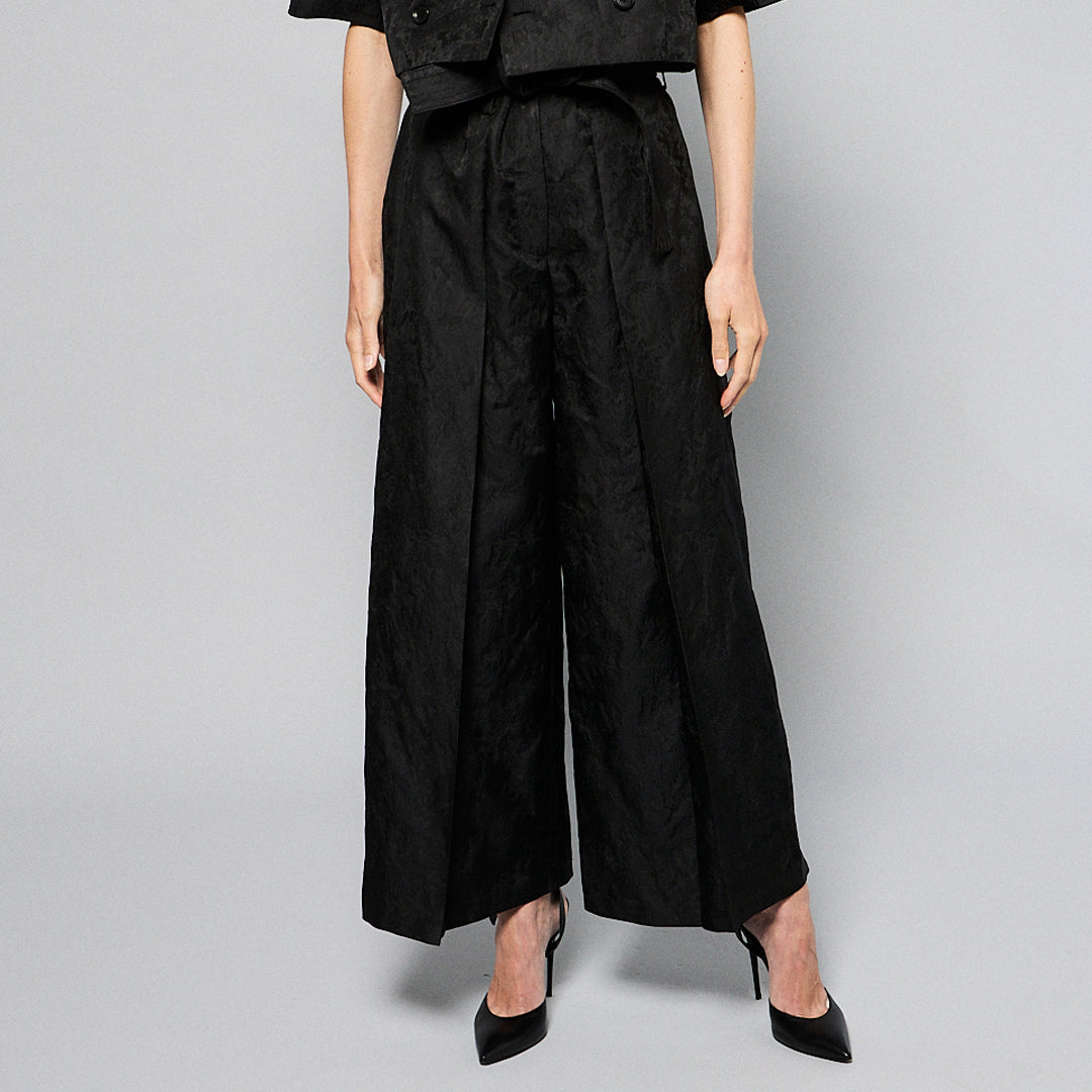  woman stands against a plain gray background, wearing a black, textured two-piece outfit with wide-leg, high-waisted Katia Pleated Hakama Pants by Maison Lilli and a short-sleeve, cropped blazer. She has her hair up and is wearing black high heels and drop earrings. A detachable belt cinches her waist.