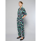 A person is wearing Maison Lilli Paperbag Pleated Pants - Floral Print with large blue, green, and white flowers. The pants have pockets and a removable belt. The person also sports a matching top and black high-heeled shoes against a plain light gray background.