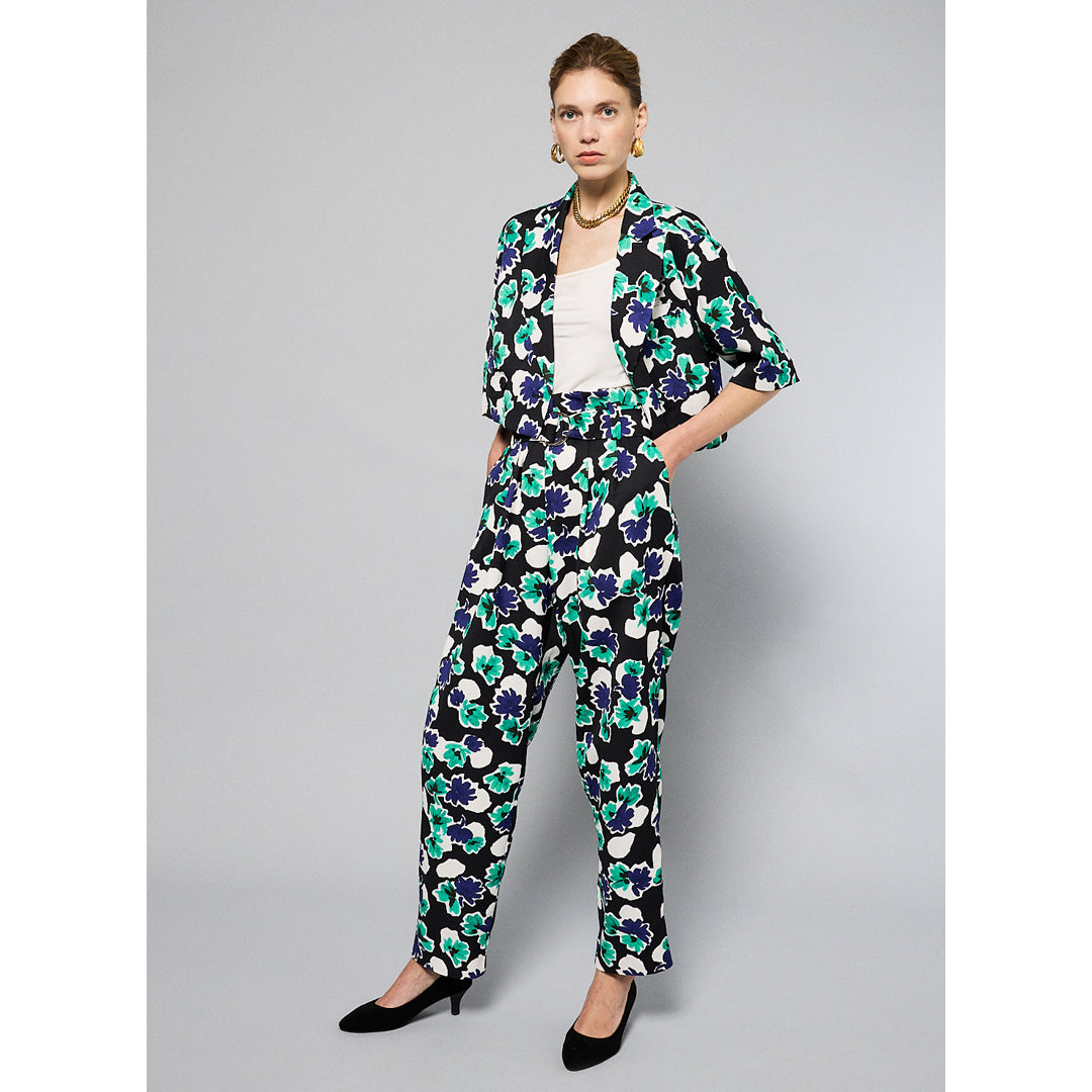 A person stands against a plain background, wearing a bucket hat and matching Yelena Short Sleeve Cropped Jacket - Floral Print by Maison Lilli in blue, green, and white. They also wear a white top underneath, gold hoop earrings, and a thick gold chain necklace.