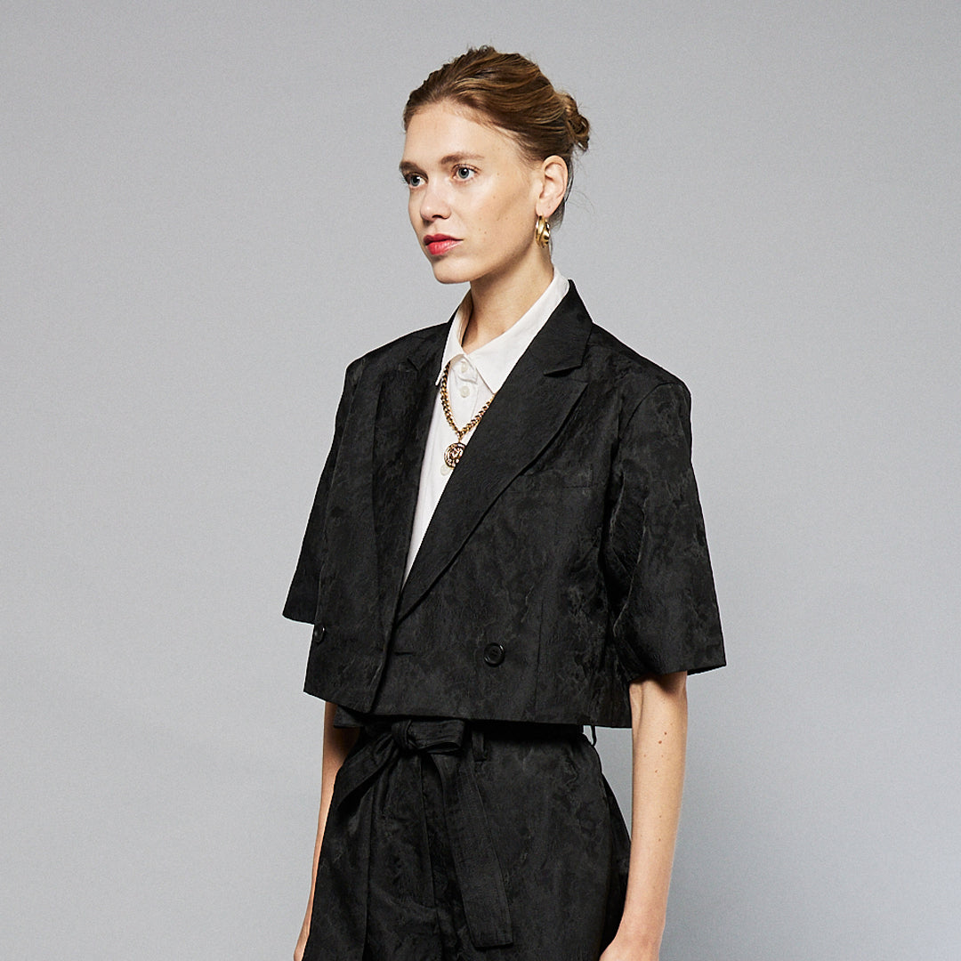 A person stands against a plain background, wearing a black Jacquard Short Sleeve Cropped Jacket  by Maison Lilli. She also wear a white top underneath, gold hoop earrings, and a thick gold chain necklace.