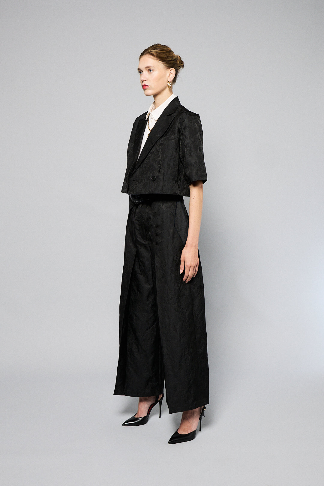 A woman stands against a plain gray background, wearing a black, textured two-piece outfit with wide-leg, high-waisted Katia Pleated Hakama Pants by Maison Lilli and a short-sleeve, cropped blazer. She has her hair up and is wearing black high heels and drop earrings. A detachable belt cinches her waist.