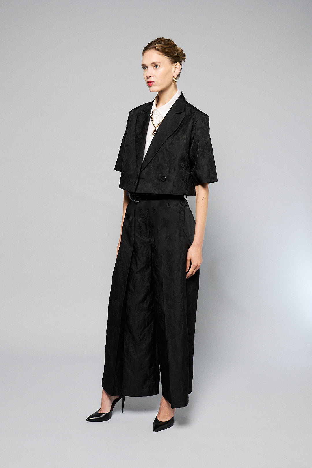  woman stands against a plain gray background, wearing a black, textured two-piece outfit with wide-leg, high-waisted Katia Pleated Hakama Pants by Maison Lilli and a short-sleeve, cropped blazer. She has her hair up and is wearing black high heels and drop earrings. A detachable belt cinches her waist.