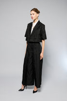  woman stands against a plain gray background, wearing a black, textured two-piece outfit with wide-leg, high-waisted Katia Pleated Hakama Pants by Maison Lilli and a short-sleeve, cropped blazer. She has her hair up and is wearing black high heels and drop earrings. A detachable belt cinches her waist.