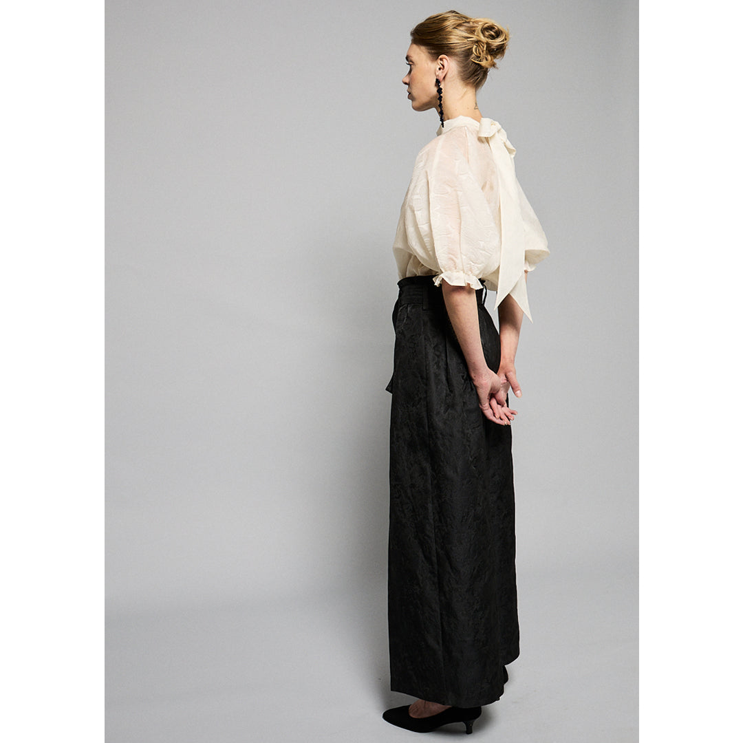  woman stands against a plain gray background, wearing a black, textured two-piece outfit with wide-leg, high-waisted Katia Pleated Hakama Pants by Maison Lilli and a short-sleeve, cropped blazer. She has her hair up and is wearing black high heels and drop earrings. A detachable belt cinches her waist.