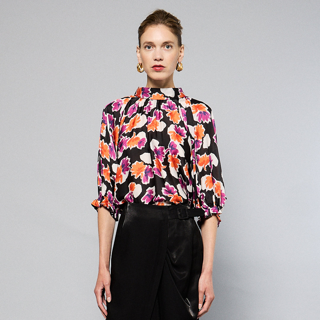 A woman wearing a Maison Lilli Amelie Floral Printed Top with vibrant pink and orange flowers and black pants stands against a grey background. Her hair is styled back, and she wears large, round earrings.