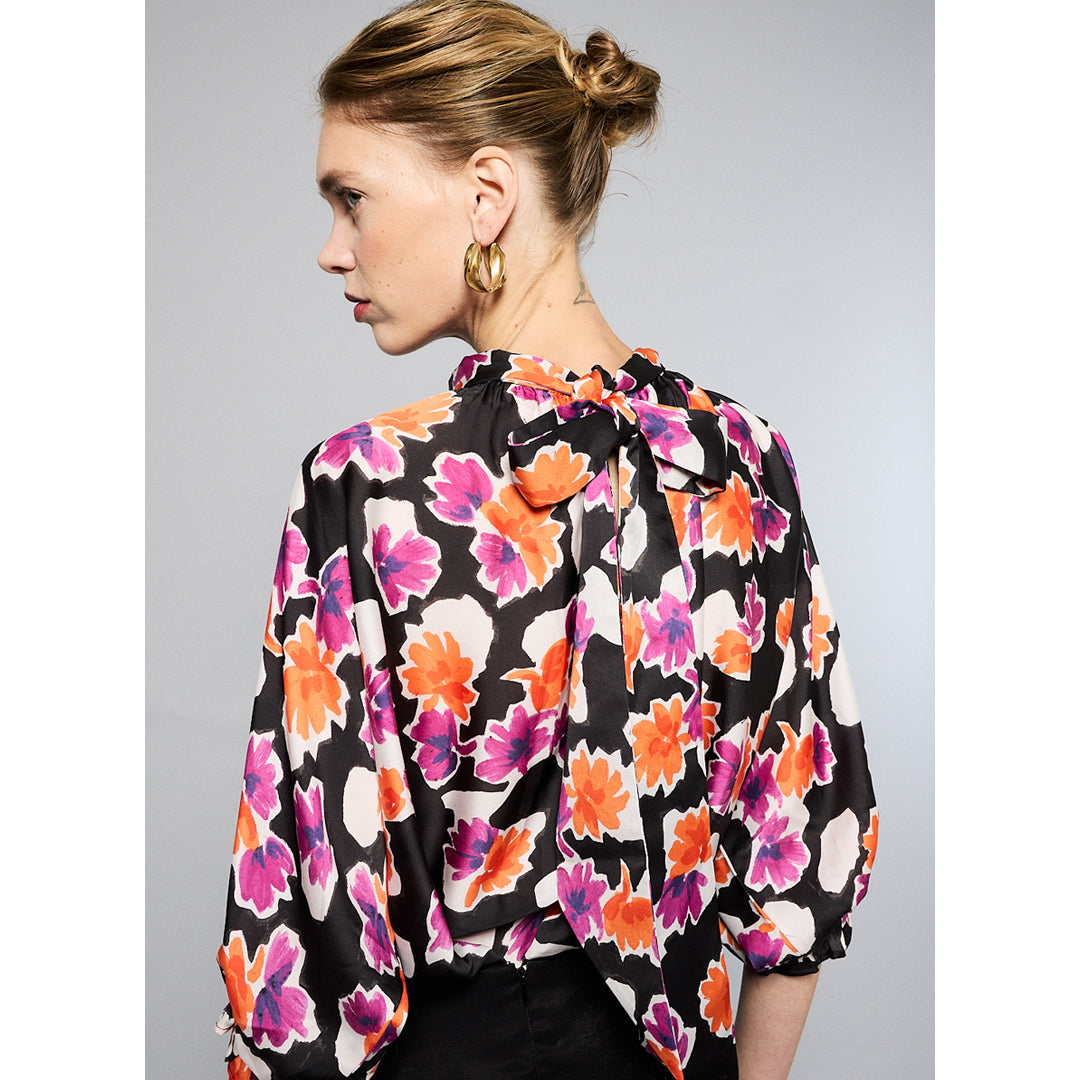 A woman wearing a Maison Lilli Amelie Floral Printed Top with vibrant pink and orange flowers and black pants stands against a grey background. Her hair is styled back, and she wears large, round earrings.